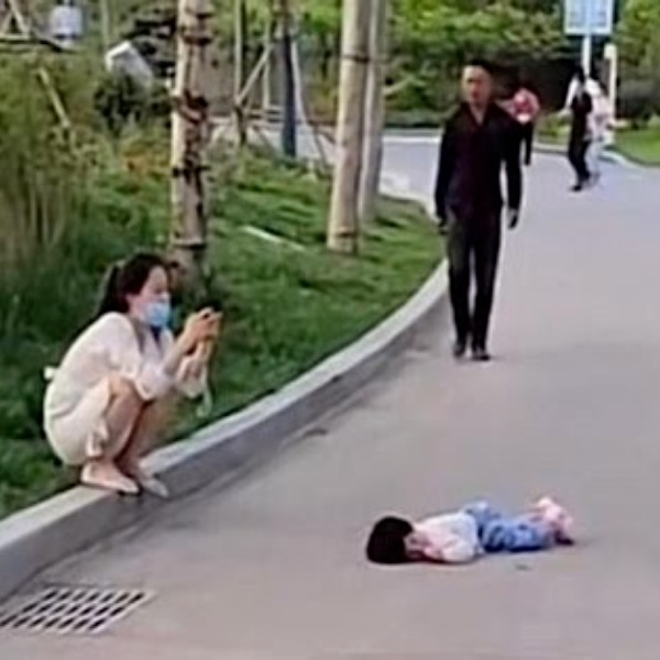  Người mẹ thản nhiên ngồi bấm điện thoại mặc kệ con nằm vạ giữa đường. (Ảnh: Sohu)
