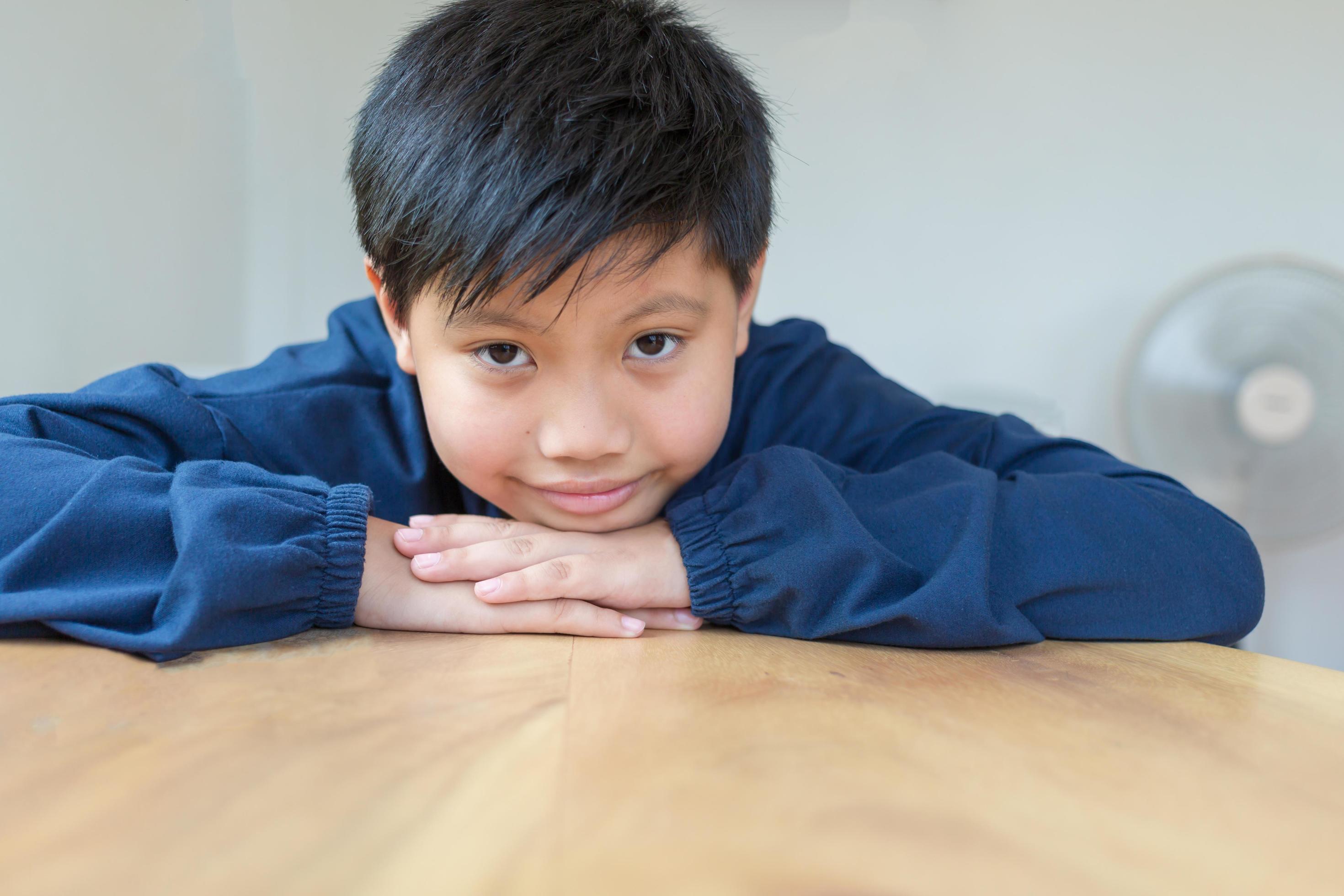 cute asian boy with white skin smiling looking in camera happily resting chins on hands on wooden table close up portrait of a cute child free photo